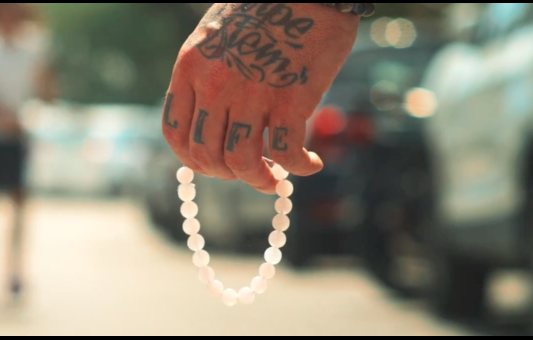 A tattooed hand holding a white beaded bracelet, with la vie and LIFE inked on the fingers, stands out against a blurred urban street scene.